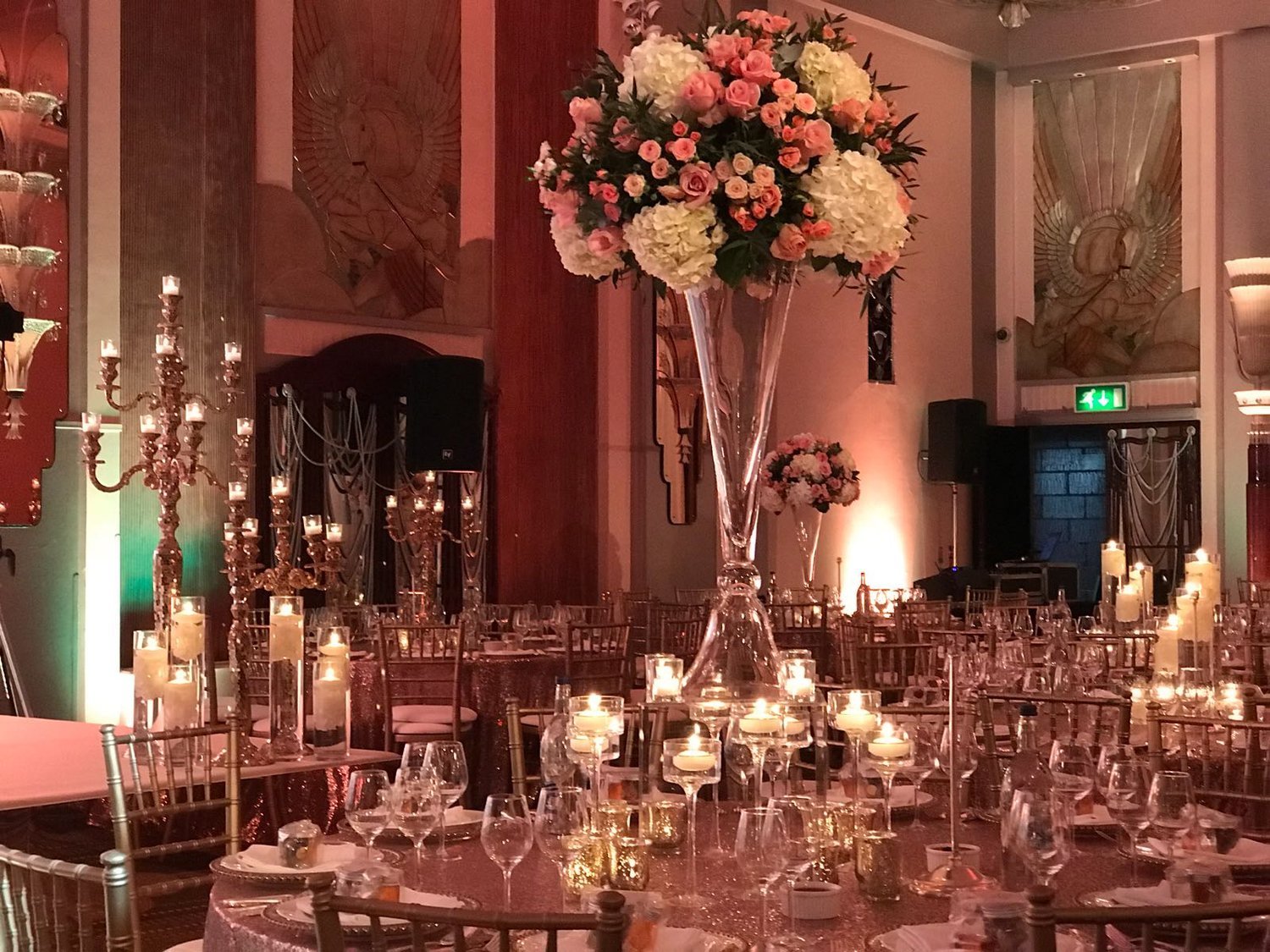 A tall glass vase of pink and ivory roses rising above a candlelit round table in an Art Deco ballroom