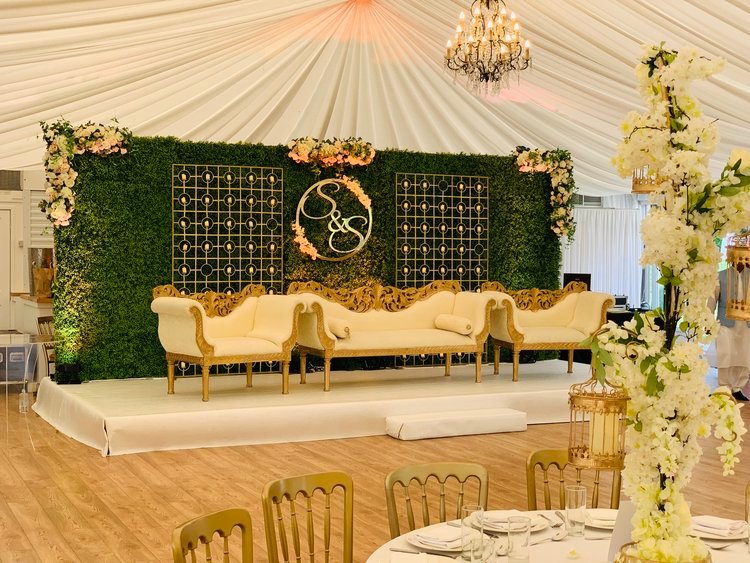 A raised reception stage with a living greenery wall, gold monogram and ivory sofas inside a draped marquee