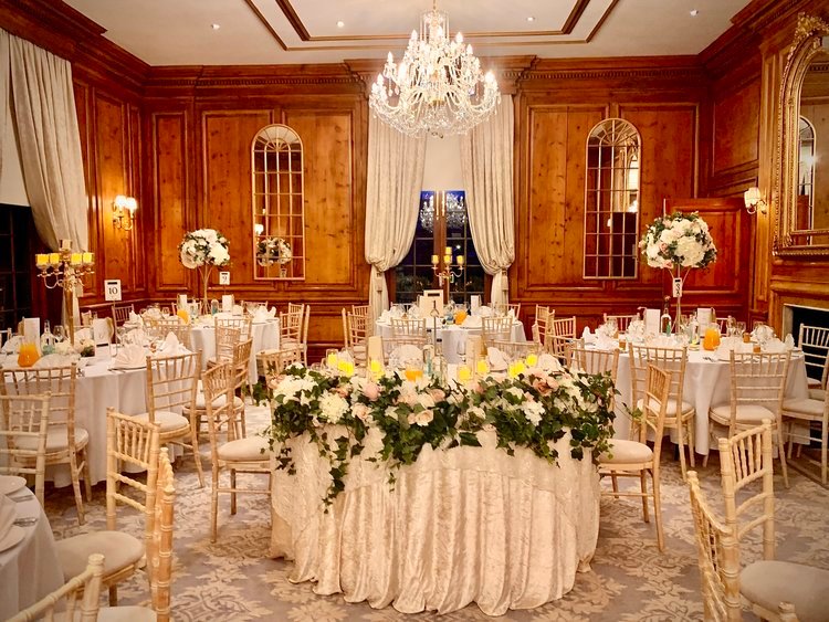 A wood-panelled reception room with ivory-draped tables, a floral-garlanded sweetheart table and a crystal chandelier