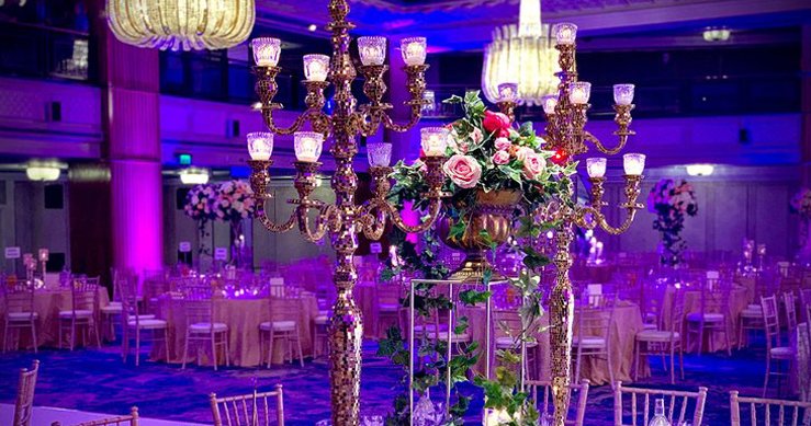 A ballroom washed in violet uplighting, with tall candelabra centrepieces of pink roses above round tables