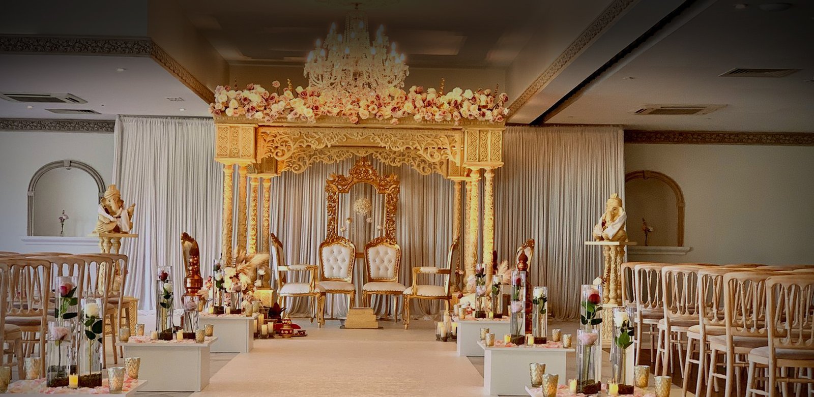 A carved gold mandap crowned with blush roses, set between ivory drapes with an aisle of floral pedestals