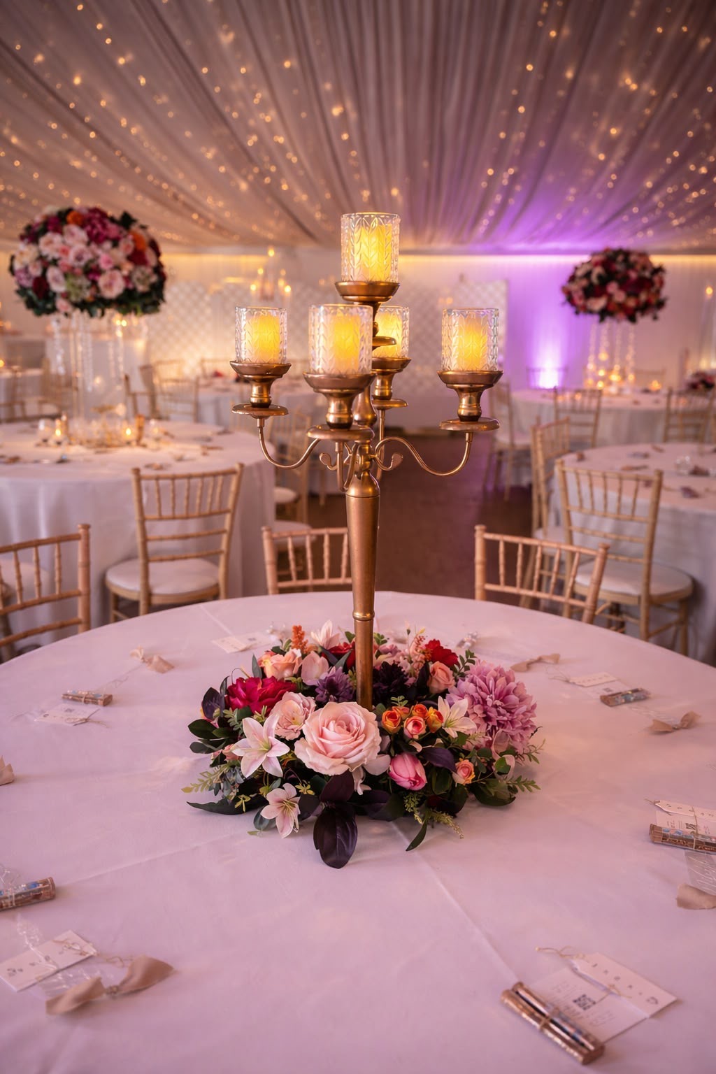 A gold five-arm candelabra rising from a wreath of pink roses, lilies and dahlias on a round table in a fairy-lit marquee
