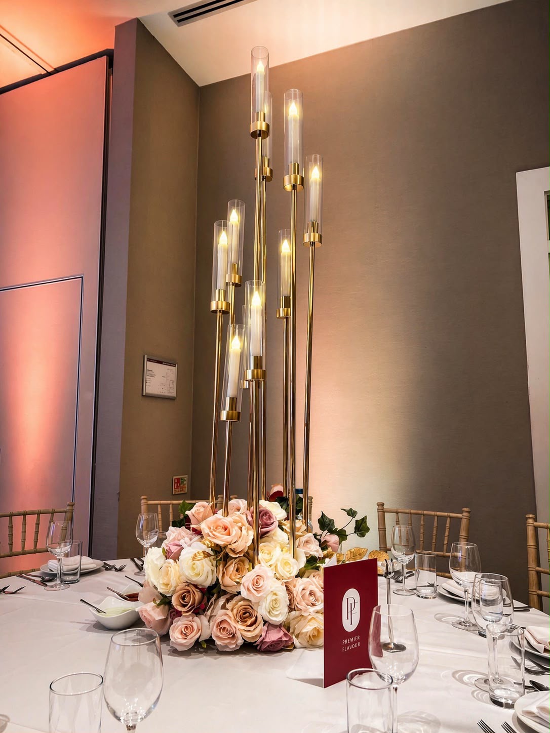 A tall gold candle-tower centrepiece with glass-shaded candles rising from a bed of peach, blush and ivory roses