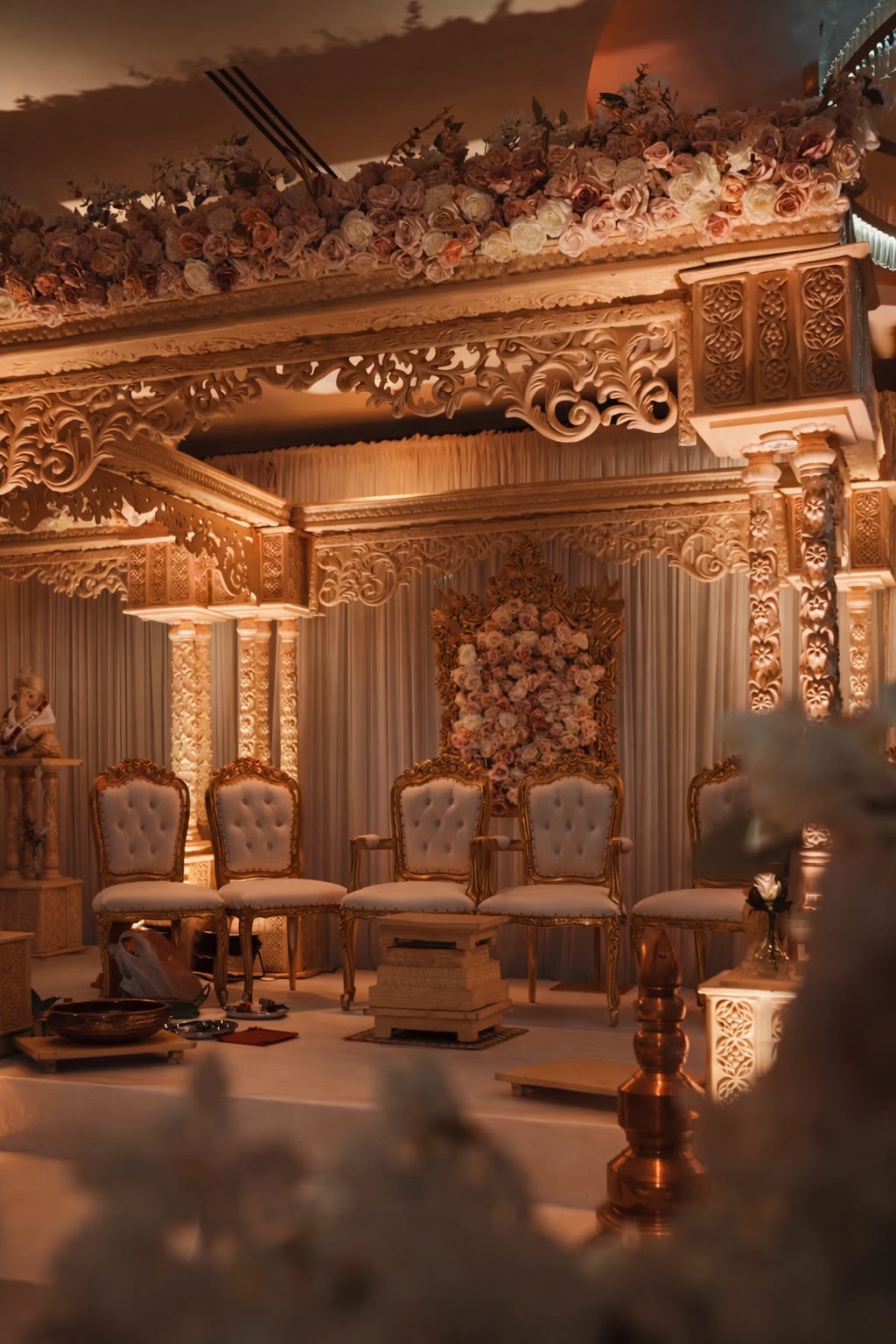 A carved ivory mandap glowing in warm light, with blush roses along its canopy and tufted gold chairs beneath