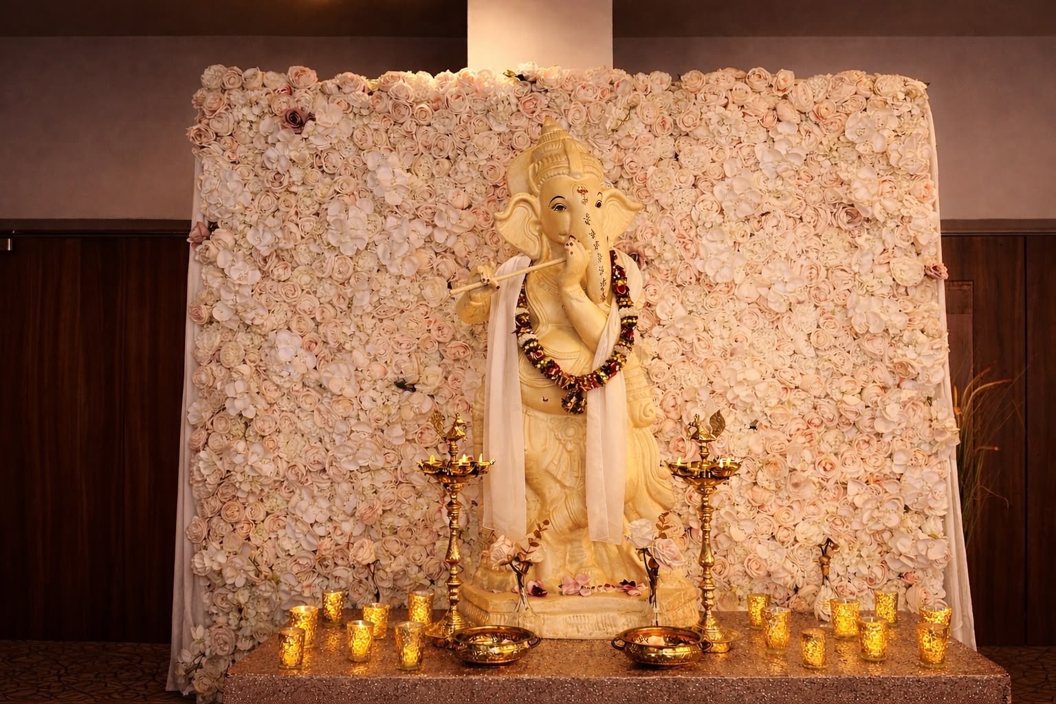A Ganesh statue garlanded with marigolds before a blush rose and orchid flower wall, with brass diyas and gold candle holders
