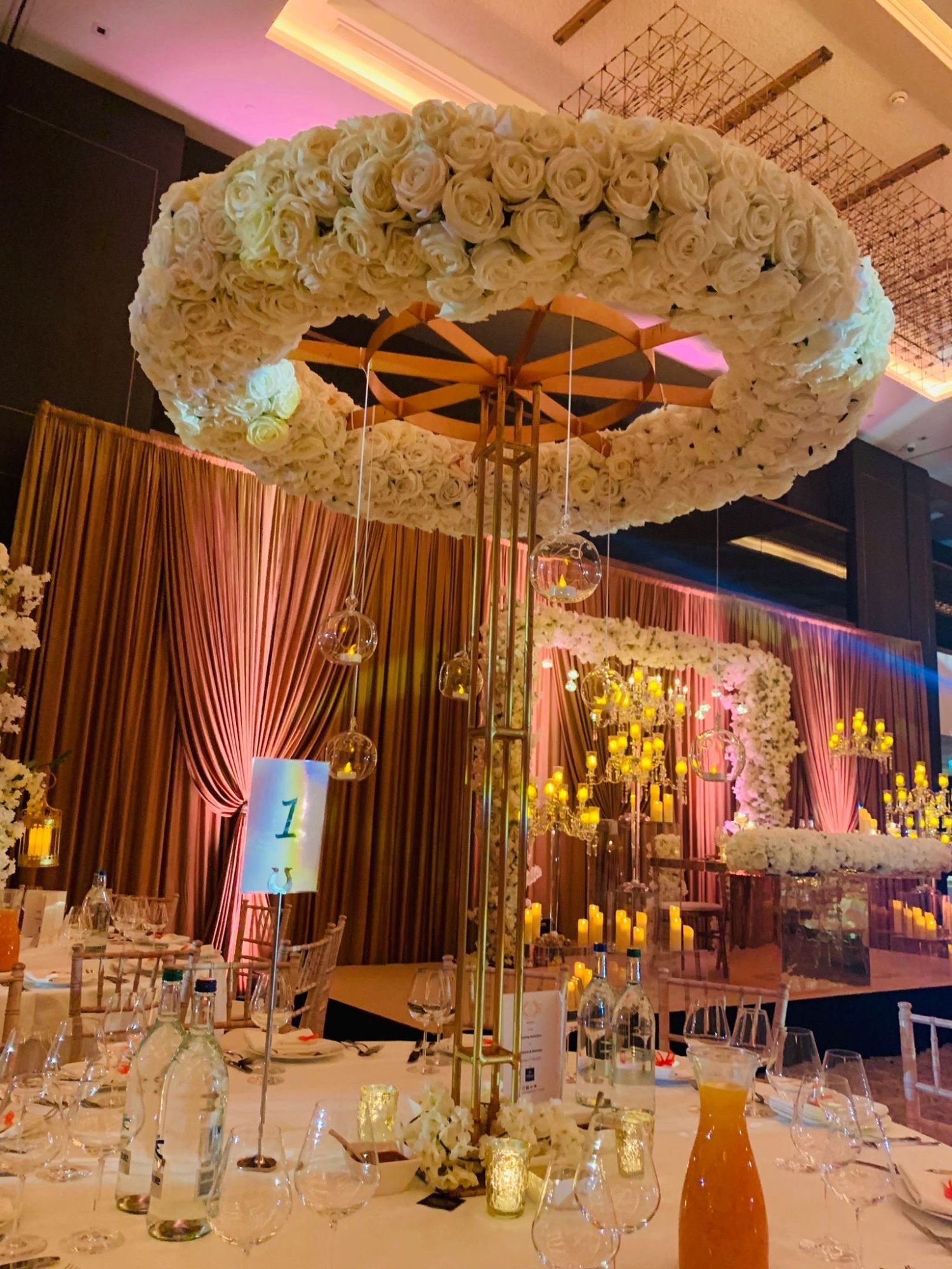 A suspended ring of ivory roses above a gold column with hanging glass tealights, with a candlelit top table and blush drapes beyond