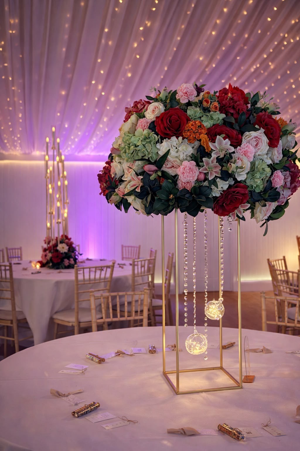 A gold stand centrepiece of red roses, pink carnations, lilies and green hydrangea with hanging crystals, in a fairy-lit marquee