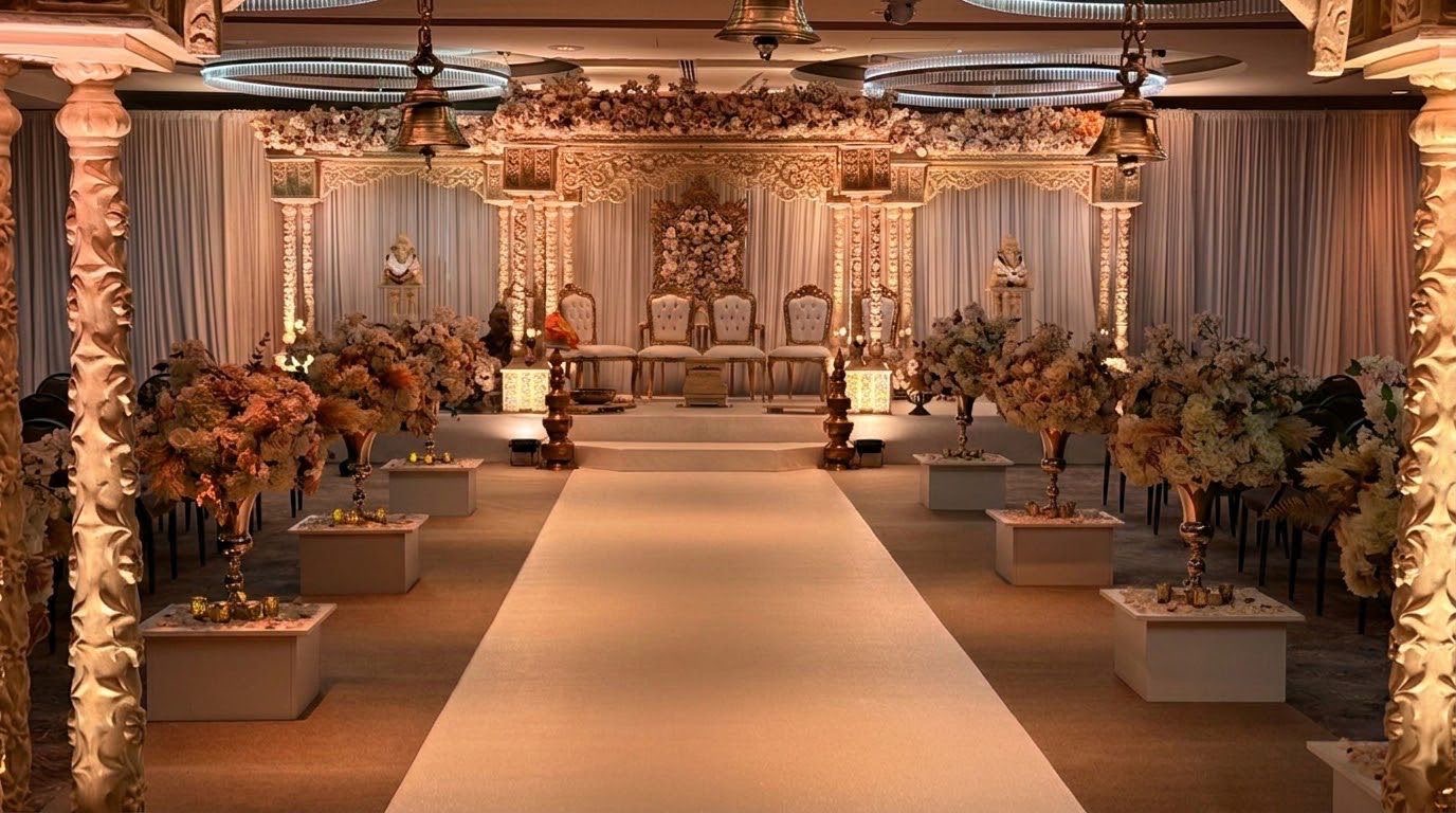 A carved ivory mandap at the end of a long aisle lined with floral pedestals, beneath hanging brass bells and halo chandeliers