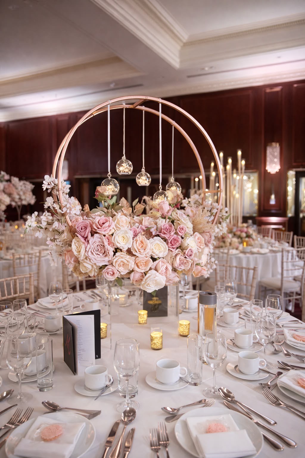 A rose-gold hoop centrepiece of blush and ivory roses with suspended glass tealight globes, above a laid table in a panelled ballroom